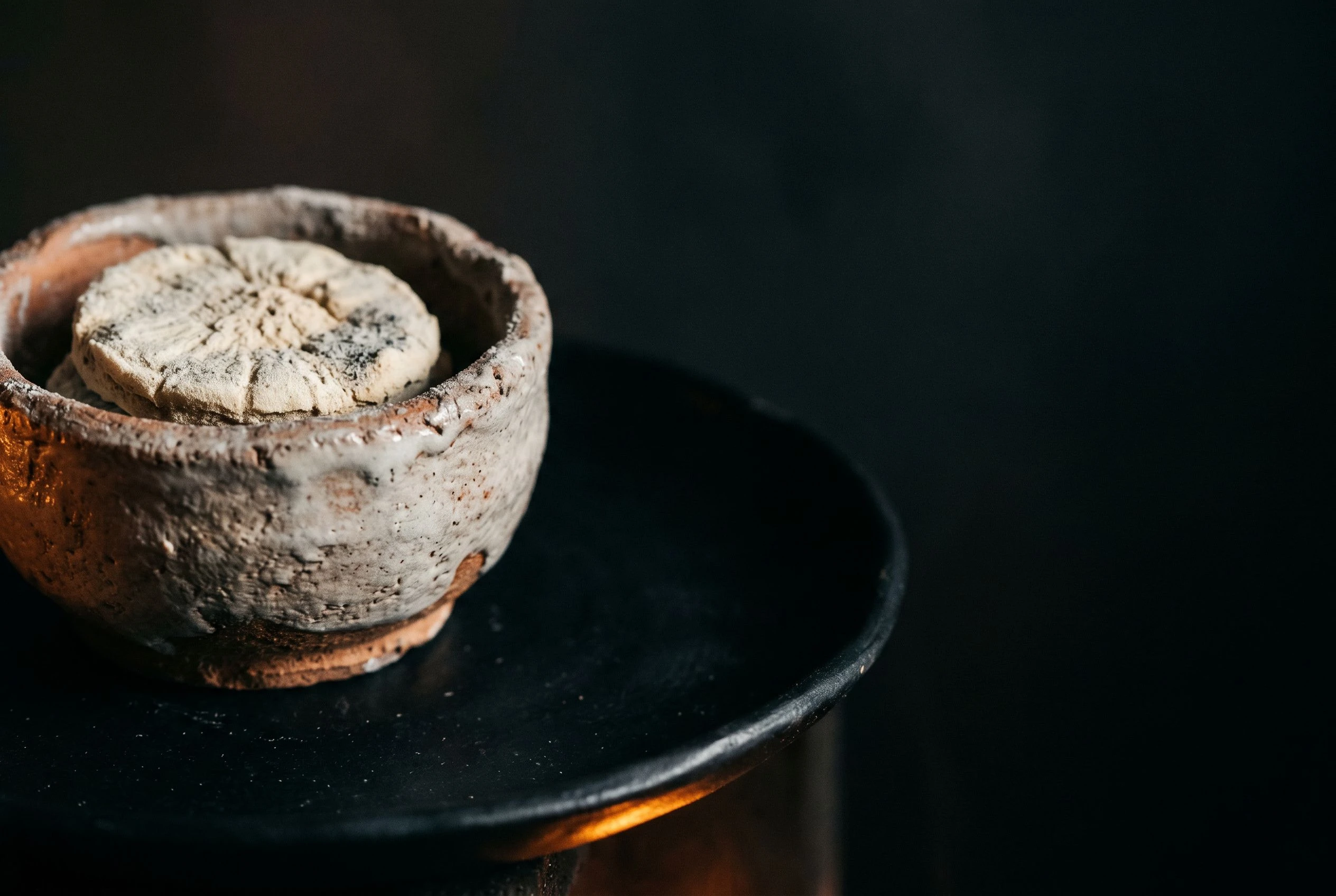 A hand-thrown ceramic vessel holding a baobab preparation, coated in pale ash glaze, resting on an obsidian-black plate in amber firelight
