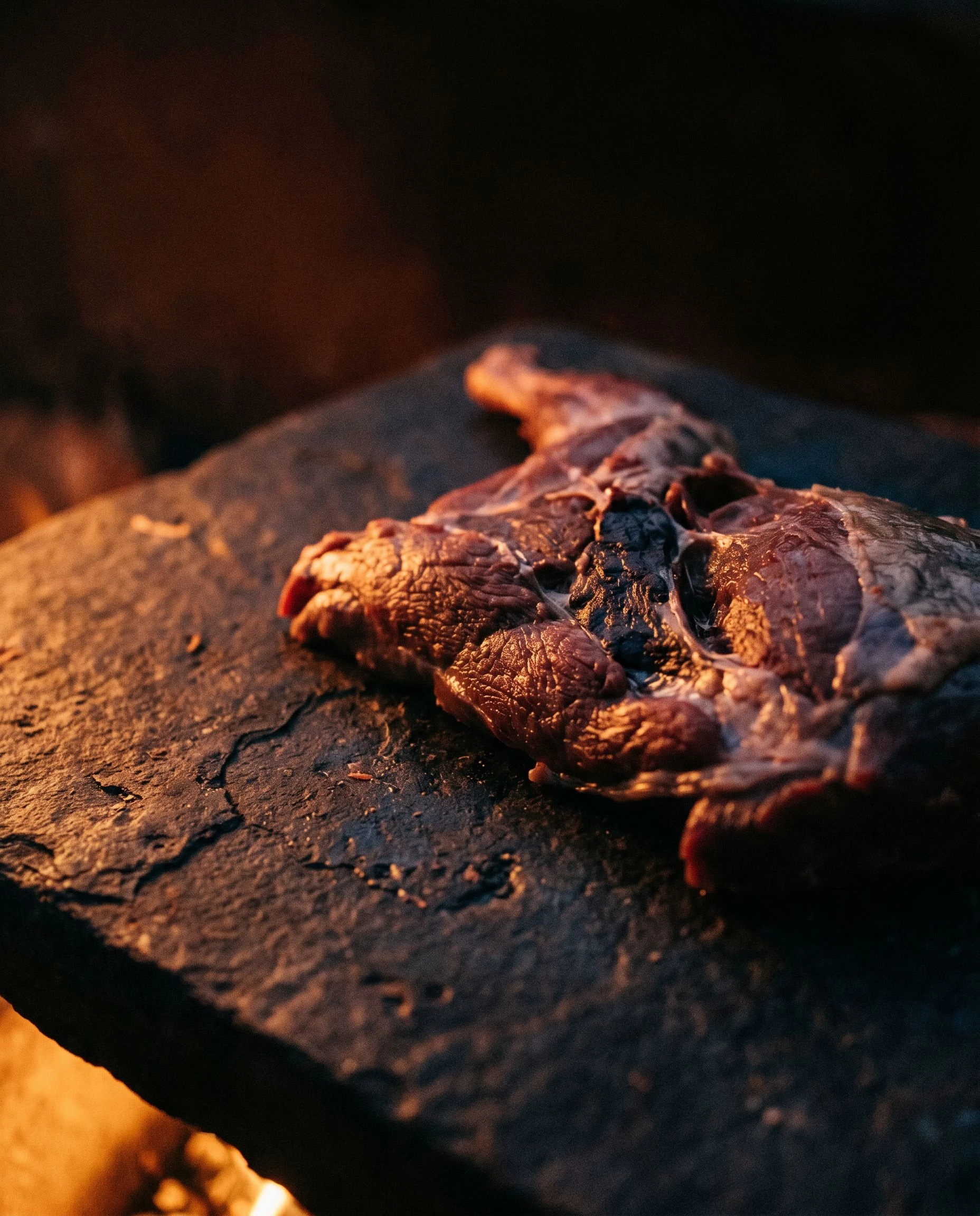 Raw wild game meat on a rough dark stone surface, firelight from below illuminating the surface, artisan butchery composition