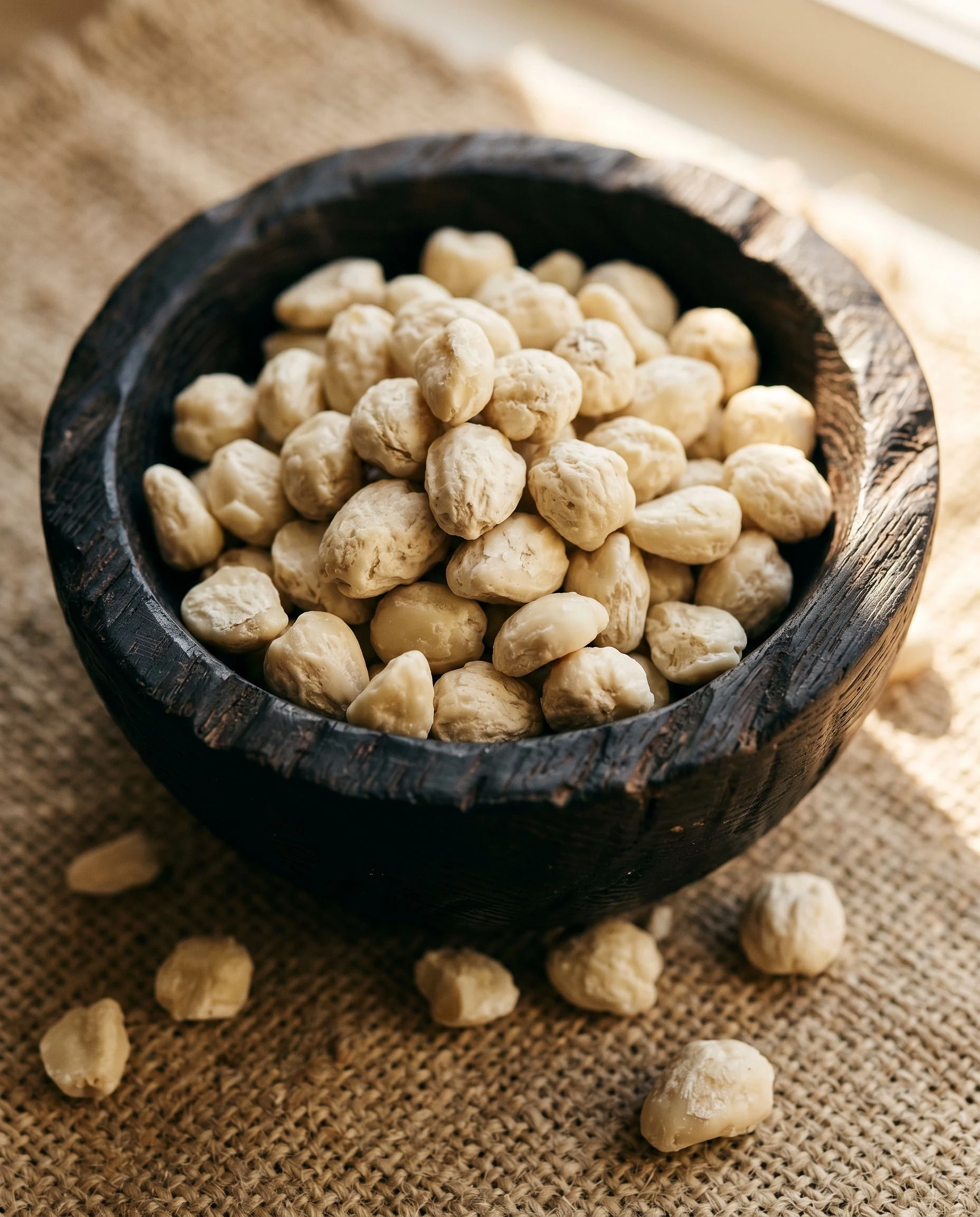 Pale shea kernels heaped in a dark carved wooden bowl, resting on a rough natural-weave textile, amber side light