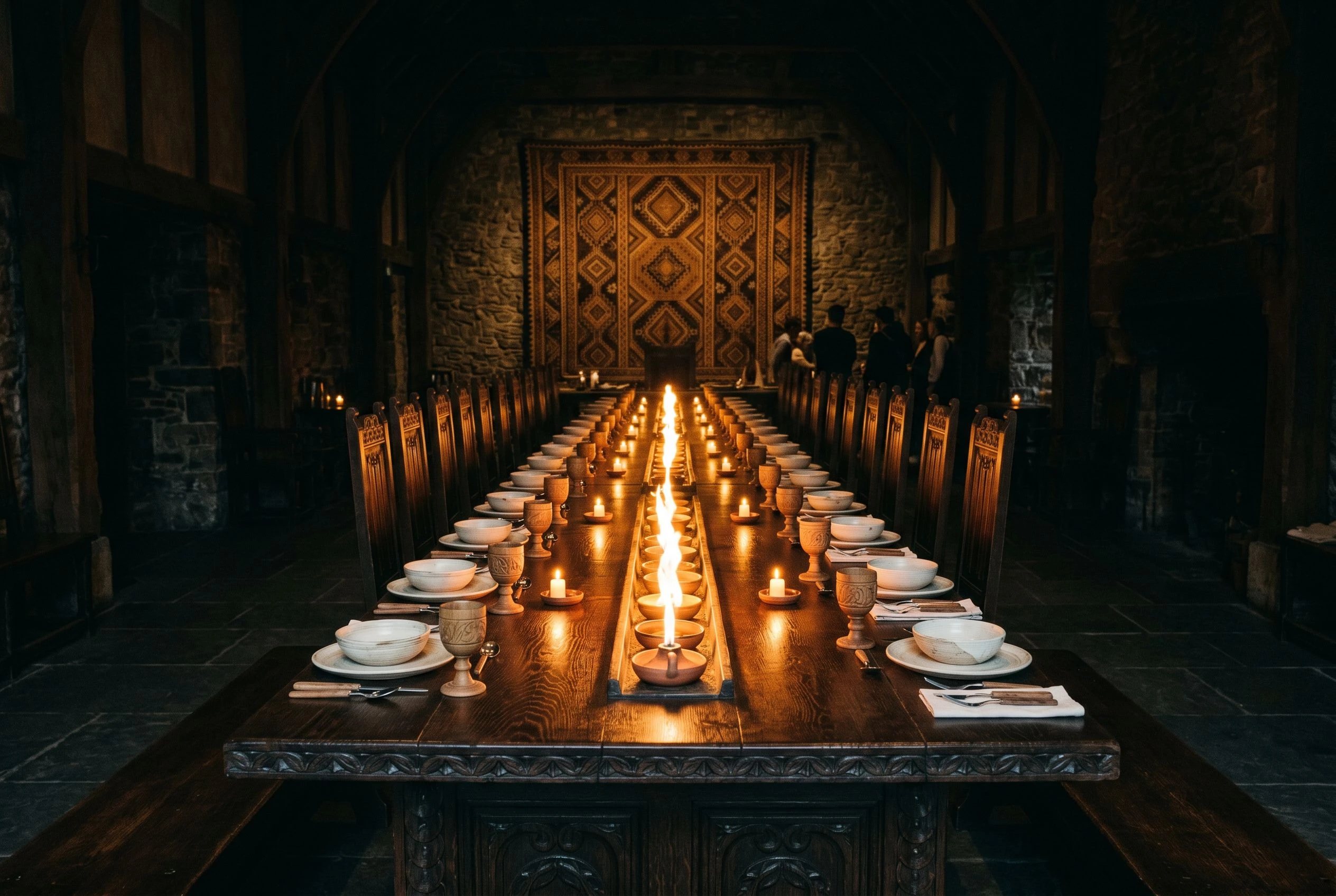 A long private dining table set for an intimate event, tribal textile backdrop, fire element centrepiece, amber light across dark carved surfaces